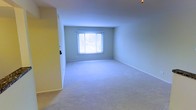 This single-floor space presents a straightforward, functional layout centered around a generously sized carpeted room. The room is finished with light-colored, wall-to-wall carpeting that extends throughout, providing a soft underfoot feel and contributing to a quiet, comfortable atmosphere. The walls are painted in a neutral, light tone, creating a bright and open ambiance that makes the space feel airy and welcoming. A centrally positioned window fitted with horizontal blinds allows natural light to filter into the room, illuminating the interior while offering adjustable privacy. The window also provides a view to the exterior surroundings. A doorway is visible to one side of the room, indicating access to adjacent areas of the space. White baseboards run along the perimeter of the walls, adding a clean, finished look to the room. The ceiling is smooth and unadorned, maintaining a simple aesthetic throughout. Overall, the space offers a clean, move-in ready environment with a neutral palette that provides versatility for a variety of uses and furnishing styles.
