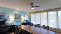 This single-story space features a thoughtfully designed conference room that balances professionalism with comfort. The room centers around a large, dark-stained oval conference table surrounded by cushioned black leather chairs, providing ample seating for meetings and collaborative sessions. A modern ceiling fan with an integrated light fixture is mounted overhead, complementing the recessed lighting to ensure consistent, well-distributed illumination throughout the space.

The walls feature a dual-tone color scheme, with a calming teal-blue accent wall adding visual interest alongside a warm neutral tone on the adjacent walls. A framed landscape painting adorns the teal wall, contributing an artistic touch, while a round decorative mirror adds depth to the space. A dedicated workstation area with a desk, monitor, and printer occupies one corner, offering functional support for presentations or administrative tasks.

Along the far wall, a row of large windows dressed with sheer roller shades allows generous natural light to filter in while maintaining privacy. Views of outdoor greenery are visible through the windows, creating a pleasant, open ambiance that enhances the overall working environment.