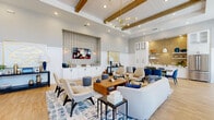 Located in Kissimmee, Florida, this thoughtfully designed single-floor community clubhouse offers a generously sized gathering space that blends modern aesthetics with warm, inviting details. The open-plan interior is anchored by soaring ceilings adorned with exposed natural wood beams and white shiplap panels, creating a coastal-contemporary ambiance throughout. A striking mid-century modern chandelier with brass accents serves as a focal point, complementing the light hardwood flooring that runs seamlessly across the space. The main lounge area features a curated seating arrangement with neutral sofas, accent chairs, and a bold blue-and-white patterned area rug. A built-in media wall with crisp white cabinetry and patterned wallpaper adds visual interest. Floor-to-ceiling windows along one wall flood the interior with natural light and provide views of the outdoor amenity area. A dedicated dining nook with a round table and yellow upholstered chairs offers a casual gathering spot, separated by a decorative frosted-glass door. The kitchenette at the rear showcases gold tile backsplash, white cabinetry, open shelving, and stainless steel appliances. White wainscoting throughout unifies the space with a clean, polished finish.