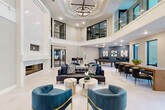 Located at 101 Crawford St in Houston, TX, this elegantly appointed communal lounge space offers a refined atmosphere ideal for socializing and relaxation. The centerpiece of the room is a dramatic double-height ceiling with a coffered tray detail, from which a striking circular chandelier descends, casting warm light throughout the open-plan interior. Large black-framed glass doors and windows line the perimeter, inviting abundant natural light and providing views of the surrounding area.

A floor-to-ceiling white marble fireplace anchors one wall, creating a luxurious focal point complemented by plush tufted leather sofas and curved teal velvet accent chairs with gold-toned legs. Light travertine tile flooring runs seamlessly throughout, enhancing the sense of openness and continuity. A mezzanine-level overlook with a glass railing adds architectural depth to the space.

The lounge transitions into a games area featuring a full-size billiards table and sophisticated dining seating, perfect for entertaining. A separate wet bar area showcases an expansive white marble countertop with bar seating, flanked by rich blue accent walls and contemporary wall sconces. Behind the bar, stainless steel appliances and ample cabinetry provide full service functionality. The overall design palette of crisp whites, cool blues, and warm metallics creates a cohesive, upscale ambiance throughout.