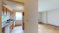 This single-floor residence at Madison Gardens offers a practical and comfortable layout centered around clean, bright interiors. The kitchen is equipped with a gas range and full-sized refrigerator, complemented by warm wood-toned cabinetry. A window above the counter invites natural light and frames a view of surrounding greenery, creating an airy atmosphere. Light-toned hardwood flooring runs throughout the space, lending warmth and visual continuity from room to room. The adjacent living area is open and unobstructed, with white walls that reflect light and contribute to a clean, neutral aesthetic. A baseboard heating system is present in both the kitchen and living areas, ensuring year-round comfort. The entryway is clearly defined, with a solid white door providing a straightforward point of entry. The overall design is understated and functional, with an emphasis on usability and natural light. The layout flows efficiently between spaces, making the home feel cohesive and well-organized. Madison Gardens provides a welcoming residential setting suited to a range of lifestyles.