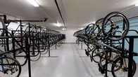 This single-floor indoor bicycle storage facility is purpose-built to accommodate a large volume of bicycles in an organized and efficient manner. The space is defined by long, parallel rows of heavy-duty black metal bike racks that run the full length of the room, creating a clear central aisle for easy access and navigation. The racks are designed to hold bicycles at alternating heights — with some bikes stored at ground level and others elevated — maximizing the use of vertical space and significantly increasing overall storage capacity. The flooring is a smooth, light-colored sealed surface that is easy to maintain and provides a clean, functional environment. The ceiling is finished in white and fitted with recessed fluorescent lighting panels that ensure the entire space is well-lit throughout. The walls are clean and white, contributing to a bright and orderly atmosphere. The layout is methodical and practical, clearly designed with high-volume cycling infrastructure in mind, making this facility well-suited for residential buildings, transit hubs, offices, or other high-traffic destinations where secure, sheltered bicycle storage is a priority.