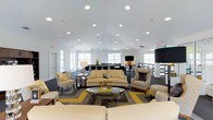 This single-floor space offers a spacious, open-concept layout designed for both comfort and functionality. The room is anchored by soaring vaulted ceilings fitted with an array of recessed lighting, ensuring the interior remains well-lit throughout. Pendant lights hang gracefully above the central seating area, adding a refined touch to the overhead design.

The lounge area is thoughtfully furnished with a neutral-toned sofa and coordinating armchairs arranged around a sleek glass coffee table. A bold geometric area rug in grey and yellow grounds the seating arrangement and introduces a vibrant accent color that carries through the décor. Additional accent chairs are positioned around the perimeter, offering flexible seating options.

Large windows line the walls, drawing in generous natural light and contributing to the room's airy atmosphere. Toward the rear of the space, a workstation or reception area is visible, suggesting a multifunctional use. A large flat-screen display is mounted nearby, making the space well-suited for presentations or community gatherings. The overall ambiance is modern, welcoming, and professionally appointed.