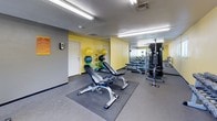 This single-level fitness center is designed to support a well-rounded workout routine, offering a comprehensive range of equipment within a functional and organized layout. The walls are finished in a bright yellow and gray color scheme, contributing an energetic atmosphere, while fluorescent lighting ensures the entire space is well-illuminated throughout.

The strength training area is a central highlight, featuring an adjustable weight bench, a multi-station cable machine, and a full dumbbell rack with a variety of weight options. A large wall-mounted mirror spans a significant portion of the room, visually expanding the space and aiding users during exercise. A dedicated medicine ball rack displays an assortment of weighted balls in multiple sizes, supporting functional fitness and core training.

The cardio section is equipped with a treadmill and an elliptical machine, both positioned near windows that allow natural light to filter into the space. A wall-mounted flat-screen television is installed nearby, providing entertainment during cardio sessions.

Rubber floor mats are placed beneath the strength equipment to protect flooring and provide stability. The space also includes a keypad entry system and clearly marked exit signage, reflecting a practical and safety-conscious design.