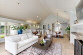 Located in Charlotte, NC, this single-floor community space offers an open, airy layout that seamlessly connects the living and dining areas. The soaring vaulted ceilings are a defining architectural feature, amplifying the sense of spaciousness throughout. Light wood flooring runs continuously across the space, complementing the soft, neutral color palette of warm whites and muted grays.

The living area is anchored by a striking stone fireplace with a white mantel and flanked by built-in cabinetry, providing both function and visual interest. A mounted television sits above the mantel, while a set of tall arched windows and French doors flood the space with natural light and offer direct views of an outdoor pool area, creating a seamless indoor-outdoor connection.

The dining area features multiple round tables and upholstered seating beneath an elegant chandelier, all set against the backdrop of the dramatic vaulted ceiling. A peninsula counter with bar-height seating and white cabinetry defines a service area at the rear, accented by recessed lighting. Crown molding and thoughtfully placed accent lighting add refined detail throughout. Together, these elements create an inviting, well-appointed gathering space suited for community use and social occasions.