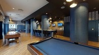 The club room at 255 Huguenot St in New Rochelle, NY, is a thoughtfully designed amenity space that combines contemporary style with a range of entertainment options. The room is anchored by warm hardwood flooring and defined by striking cylindrical columns wrapped in textured blue tile, which serve as both structural and decorative elements throughout the open layout.

A full-size billiards table takes center stage in one section of the room, positioned beneath an industrial-style pendant light fixture and flanked by large windows that allow natural light to filter in and offer views of the surrounding streetscape. Adjacent to this area is a ping-pong table, providing additional recreational options within the same open-plan space.

The lounge area features a long bar-height counter with tufted black swivel chairs, accompanied by pendant lighting and wall-mounted flat-screen televisions. Reclaimed wood accent walls and geometric-patterned wallpaper add visual texture, while the dark ceiling reinforces the room's sophisticated, moody atmosphere.

A dedicated seating nook offers a built-in upholstered sectional sofa adorned with mixed-pattern pillows, set beneath a gallery wall of framed locomotive blueprint prints — a subtle nod to the area's transit history. A separate lounge corner features a classic armchair, rounding out the variety of social and relaxation zones. The room's cohesive navy, charcoal, and natural wood palette gives the entire space a polished, urban character.
