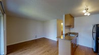 This single-floor residence is designed with an open-concept layout that seamlessly connects the living area and kitchen, creating a spacious and functional environment. The kitchen is a standout feature, boasting warm wood cabinetry, granite countertops, and a peninsula with an undermount sink — offering both ample prep space and a natural divider between the cooking and living zones. The flooring throughout is a warm-toned hardwood-style material that adds continuity and a welcoming feel to the space. Neutral-toned walls provide a clean, versatile backdrop that complements a variety of interior styles. A textured ceiling runs throughout the unit, adding subtle visual interest overhead. A floor-level vent indicates a central heating system, contributing to year-round comfort. A hallway visible in the background leads to additional areas of the unit, hinting at a practical and well-organized floor plan. The overall ambiance is bright, clean, and move-in ready, with a balanced blend of modern finishes and functional design elements that make it a comfortable and adaptable living space.