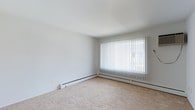 This single-floor space presents a straightforward, functional layout centered around a generously sized room with a clean, minimalist aesthetic. The walls are finished in crisp white, creating a bright and open atmosphere that serves as a blank canvas for a variety of interior design styles. Underfoot, wall-to-wall beige carpeting adds warmth and comfort to the space. A large picture window dominates one wall, dressed with vertical blinds that allow for adjustable natural light control. The window draws in ample daylight, contributing to the room's airy feel. Along the base of the walls, a continuous baseboard heating system ensures efficient and even climate control throughout the space. The smooth white ceiling complements the neutral palette, maintaining a cohesive and uncluttered look. The overall ambiance is calm and understated, well-suited for those seeking a clean, move-in-ready environment with practical features and a versatile foundation for personalization.