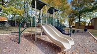 Located on Caroline Court in Circleville, Ohio, this outdoor recreational space offers a well-equipped playground area designed for active play and community enjoyment. The centerpiece of the space is a multi-level play structure finished in green and grey, featuring dual side-by-side slides, a covered upper platform, climbing stairs with handrails, and various interactive elements. The structure is set within a wood chip safety surface that defines the play zone and provides impact cushioning underfoot. Surrounding the playground, a spacious open lawn extends outward, bordered by a wooden privacy fence that encloses the area and provides a sense of security. Mature deciduous trees are distributed throughout the grounds, offering natural shade and a parklike atmosphere. A picnic table is positioned nearby, providing a convenient gathering spot for visitors. The open grassy areas beyond the playground offer additional room for outdoor activities. The combination of the structured play equipment, shaded green space, and fenced boundaries makes this a functional and inviting community recreational area suitable for a range of outdoor uses.