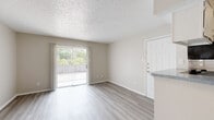 This single-story unit is designed with a clean, neutral aesthetic throughout, featuring light wood-look flooring that flows consistently across the main living areas and bedroom. The open living area is bright and functional, anchored by a sliding glass door with vertical blinds that leads to a balcony offering views of surrounding greenery, allowing natural light to fill the space. A white entry door and textured ceiling complete the living room's straightforward layout.

The kitchen is positioned adjacent to the living area and features a pass-through counter with a speckled countertop, white cabinetry, subway tile backsplash, and stainless steel appliances including a refrigerator, dishwasher, and microwave. The galley-style layout makes efficient use of the available space.

The bedroom is a well-proportioned room equipped with a ceiling fan, a window fitted with horizontal blinds for natural light and privacy, and direct access to a closet. The bathroom includes a full-size tub and shower combination with white tile surround, a toilet, and a vanity. A dedicated laundry nook accommodates a full-size top-load washer, with shelving above for additional storage. The overall ambiance is clean, practical, and move-in ready.