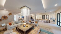 Located at 2900 N Braeswood Blvd in Houston, Texas, this elegantly designed single-floor space offers a seamless blend of comfort and contemporary sophistication. The main lounge area serves as the centerpiece, anchored by a striking cascading crystal chandelier that draws the eye upward to the crisply detailed ceiling with recessed tray lighting. Plush, neutral-toned sofas and accent chairs are arranged around a substantial wood coffee table atop a richly patterned area rug, creating an inviting and refined atmosphere. A textured tile accent wall adorned with sculptural botanical medallions adds an artistic focal point, complemented by a large wall-mounted flat-screen television nearby. The space flows openly into a formal dining area furnished with a long wooden table and upholstered chairs, bathed in natural light from expansive floor-to-ceiling black-framed glass doors and windows that frame views of the surrounding outdoor greenery. The entry corridor features polished large-format tile flooring, bold artwork, and black-framed glass doors that reinforce the modern aesthetic. Gold linear ceiling accents in the dining zone add warmth and contrast. Throughout, the design palette of soft whites, warm neutrals, and earthy orange tones creates a cohesive, welcoming environment well-suited for both social gatherings and private meetings.