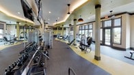 This expansive fitness center is thoughtfully designed to accommodate a full range of workout activities across a single, open floor. The space is organized into distinct zones, each tailored to a specific type of exercise, creating an efficient and intuitive layout for users.

The strength training area features a comprehensive selection of weight machines, cable systems, and a full rack of dumbbells lining one wall, accompanied by wall-mounted televisions for entertainment. Flooring transitions between charcoal gray carpet and vibrant yellow rubber matting visually delineate the different training zones. Structural columns with dark trim accents are integrated throughout, adding architectural definition without disrupting the flow of the space.

The cardio zone is positioned near floor-to-ceiling windows fitted with roller shades, allowing generous natural light and outdoor views of the surrounding landscape. Treadmills, elliptical trainers, stationary bikes, and a rowing machine are all represented, offering varied options for cardiovascular training.

A glass-enclosed cycling studio provides a semi-private space outfitted with spin bikes and mirrored walls, visible from the main floor. Pendant lighting with copper-toned shades, a coved ceiling with accent lighting, and a green upper wall treatment contribute to the modern, energetic ambiance of the overall facility.