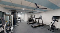Located at 4455 W New Haven Ave in Melbourne, Florida, this fitness center offers a well-equipped, single-floor workout space designed to support a comprehensive exercise routine. The layout is efficiently organized, making effective use of its footprint to accommodate a diverse range of fitness equipment without feeling cramped.

The cardio zone features multiple treadmills, an elliptical, and stationary cycling bikes — each equipped with individual display consoles for tracking workout metrics. The strength training area is outfitted with a multi-functional cable machine, a weight rack with dumbbells, a bench press, and additional resistance equipment, providing ample options for both machine-based and free weight exercises.

The interior design is clean and modern, with crisp white walls and a drop ceiling featuring teal accent tiles and recessed lighting. Large ceiling fans ensure consistent air circulation throughout the space. Full-length wall mirrors on the strength training side enhance the sense of openness and allow users to monitor their form during workouts.

Durable dark rubber flooring covers the entire space, offering both safety and comfort underfoot. A dual-basin water fountain station is conveniently positioned for easy access. The facility is accessed through frosted glass doors, contributing to the space's polished, professional aesthetic.