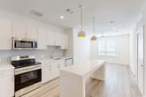 Located at 3842 W Northwest Hwy in Dallas, TX, this well-appointed one-bedroom, one-bathroom unit offers a thoughtfully designed open-concept layout finished with light wood-tone flooring throughout. The kitchen serves as the centerpiece of the living area, showcasing crisp white cabinetry, a subway tile backsplash, light quartz countertops, and a full suite of stainless steel appliances including a refrigerator with ice maker, range, microwave, and dishwasher. A large center island with seating provides additional prep and dining space, while stylish pendant lights with yellow interior accents add a modern touch overhead. The open living area benefits from natural light through a large window fitted with blinds and is equipped with a ceiling fan for added comfort. The bedroom is a calm, neutral retreat with its own ceiling fan, a sizable window, and warm wood-style flooring. The bathroom features a wide vanity with ample counter space and connects directly to a dedicated walk-in closet with built-in shelving rods. A stacked in-unit washer and dryer offer everyday convenience. Clean lines, a neutral palette, and modern finishes define the overall aesthetic of this efficient and functional space.