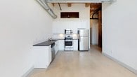 This 2-bedroom, 2-bathroom urban loft showcases a compelling blend of industrial character and contemporary design across a single floor. The kitchen is a standout space, boasting white cabinetry, dark granite countertops, and a full suite of stainless steel appliances including a refrigerator, electric range, built-in microwave, and dishwasher. Polished concrete floors run throughout, complementing the exposed metal ductwork and timber structural elements that lend the space a distinctive loft aesthetic. The soaring ceiling height adds a sense of volume and openness to the living areas, while the open-plan layout creates a seamless flow between the kitchen and main living space. Both bathrooms are finished in a clean, modern style, featuring tub-shower combinations with curved curtain rods and brushed nickel fixtures. One bathroom includes a white vanity with a dark granite countertop and undermount sink, paired with a large mirror. Light tile flooring in the bathrooms contrasts neatly with the concrete found elsewhere in the unit. Overall, the space offers a well-balanced combination of raw, industrial elements and refined modern touches, creating an inviting and functional urban living environment.