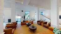 Located at 6689 El Colegio Rd in Goleta, California, this single-floor communal lobby space offers a thoughtfully designed environment that blends contemporary aesthetics with functional amenities. The entrance welcomes visitors through large glass doors framed in black, opening into an airy, high-ceilinged foyer with a chevron-patterned tile floor that transitions seamlessly into warm-toned carpeted lounge areas.

The main lounge is furnished with rich cognac leather sofas and tufted armchairs, complemented by modern round coffee tables with bowl-shaped centerpieces and accent seating. Tall black-framed windows flood the interior with natural light, offering views of the surrounding greenery. A sleek staircase runs along one wall, adding architectural interest to the open layout.

One of the most striking features is a large-scale, black-and-white line art mural spanning an entire wall, depicting abstract faces and figures that lend an artistic, creative energy to the space. Adjacent to the lounge is a dining area with round marble-top tables and leather side chairs, ideal for casual gatherings.

A dedicated mailroom provides individual resident mailboxes in a classic wood cabinet arrangement, accompanied by a beverage vending machine. Track lighting and recessed fixtures throughout ensure consistent, well-distributed illumination across all areas of this vibrant, community-centered space.