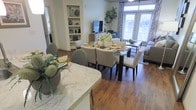 This single-floor residence showcases an open-concept layout that connects the kitchen, dining, and living areas into one cohesive, flowing space. Rich hardwood flooring extends throughout, adding warmth and continuity across the entire footprint. The kitchen features a granite-topped counter with a breakfast bar overhang, providing both prep space and a casual seating option. Adjacent to the kitchen, the dining area accommodates a full-sized rectangular table with upholstered seating for six, making it well-suited for everyday meals and entertaining. A built-in white shelving unit anchors one wall, offering structured display and storage space. The living area, situated toward the rear of the space, benefits from generous natural light streaming through large windows dressed with patterned curtains. French doors open to an outdoor view, further brightening the interior. A wall-mounted television and a sleek media console complete the living area, while a tall potted fig tree adds a striking organic element. The overall ambiance is bright, casual, and welcoming.