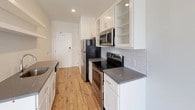 This single-floor, one-bedroom, one-bathroom unit showcases a clean, contemporary design throughout. The kitchen is efficiently laid out in a galley style, featuring white cabinetry with glass-front upper shelves, grey quartz-style countertops, white subway tile backsplash, and a full suite of stainless steel appliances including a refrigerator, electric range, and over-the-range microwave. Warm hardwood-style flooring runs throughout the kitchen and main living areas, complementing the bright, neutral color palette. A peninsula with an integrated sink and additional open floating shelves above adds functional prep and storage space, while providing a visual connection between the kitchen and entryway.

The bedroom offers plush carpeting, a ceiling fan with lighting, and a large window that fills the room with natural light and frames outdoor views. A mirrored sliding closet door leads to a well-sized reach-in closet with built-in shelving and hanging rods.

The bathroom is finished with light tile flooring, a white vanity with dark stone countertops, a large mirror, recessed lighting, and a full soaking tub with a shower combination, creating a clean and functional space.