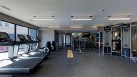 This fitness center is designed to support a comprehensive workout experience within a single, open-plan floor. The space is laid out with clear zones for different types of exercise, allowing users to move efficiently between equipment. Along one side, a row of cardio machines — including treadmills and ellipticals — lines the perimeter near large windows that bring in natural light and provide views to the exterior. Toward the center and rear of the space, a full free weights area features a well-stocked dumbbell rack alongside medicine balls and a dedicated stretching and lifting bench. Floor-to-ceiling mirrors along the back wall visually expand the space and allow users to monitor their form during training. The flooring throughout is durable rubber gym matting, suitable for heavy equipment and high-impact activity. Overhead, linear fluorescent lighting fixtures ensure consistent, bright illumination across the entire floor. The overall aesthetic is clean and modern, with a neutral color palette of grey and white that gives the space a professional, focused atmosphere. A glass-paneled entry door adds a polished finishing touch to this functional and well-appointed fitness facility.