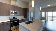This single-floor space showcases a thoughtfully designed, modern kitchen that serves as its centerpiece. The kitchen features a generous island with a light-toned quartz countertop and an undermount stainless steel sink equipped with a contemporary arc-style faucet. Two elongated pendant lights hang gracefully above, drawing the eye upward toward the notably high ceilings that lend an open, airy quality to the space.

Cabinetry is finished in rich, dark wood tones, providing a striking contrast against the light countertops and the classic white subway tile backsplash. Full-height upper and lower cabinets offer substantial storage, while stainless steel appliances—including a gas range and an over-the-range microwave—contribute to the kitchen's polished, professional aesthetic.

The flooring transitions between hardwood and other materials, adding warmth and texture throughout. Natural light filters in through a nearby window, complementing the recessed lighting overhead. The overall ambiance is clean, contemporary, and functional, with a cohesive design language carried throughout the open layout.