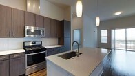 This single-floor space showcases a thoughtfully designed, modern kitchen that serves as its centerpiece. The kitchen features a generous island with a light-toned quartz countertop and an undermount stainless steel sink equipped with a contemporary arc-style faucet. Two elongated pendant lights hang gracefully above, drawing the eye upward toward the notably high ceilings that lend an open, airy quality to the space.

Cabinetry is finished in rich, dark wood tones, providing a striking contrast against the light countertops and the classic white subway tile backsplash. Full-height upper and lower cabinets offer substantial storage, while stainless steel appliances—including a gas range and an over-the-range microwave—contribute to the kitchen's polished, professional aesthetic.

The flooring transitions between hardwood and other materials, adding warmth and texture throughout. Natural light filters in through a nearby window, complementing the recessed lighting overhead. The overall ambiance is clean, contemporary, and functional, with a cohesive design language carried throughout the open layout.