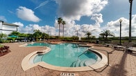 Located at 8335 Narcoossee Rd in the Lake Nona area of Orlando, Florida, this resort-style outdoor amenity complex offers a compelling blend of leisure and tropical ambiance. The centerpiece is a generously sized freeform swimming pool with crystal-clear turquoise water, complemented by a dedicated hot tub/spa area accessible via a gradual step-in entry. The pool deck is finished with warm-toned brick pavers that surround the water features, providing ample space for relaxation and recreation. Rows of chaise lounge chairs are positioned throughout the deck area, shaded in part by vibrant teal market umbrellas, making it ideal for both sunbathing and casual lounging. Tall, mature palm trees are strategically placed around the perimeter, enhancing the tropical atmosphere and providing natural shade. Lush tropical landscaping with colorful foliage and manicured greenery borders the entire space. The complex is framed by multi-story residential buildings featuring a classic white and brown architectural style with balconies overlooking the pool area. A serene waterfront view is visible beyond the fencing, adding a scenic backdrop. Ornamental lamp posts and a well-maintained iron fence complete the polished, community-focused aesthetic of this outdoor retreat.