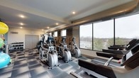 This fitness center offers a thoughtfully designed workout space that combines functionality with an appealing aesthetic. The room features a row of elliptical trainers as a centerpiece, complemented by treadmills and additional strength and cardio equipment positioned toward the back. Large floor-to-ceiling windows along one wall flood the space with natural light while providing elevated views of the surrounding landscape, creating an inviting environment for exercise. The exposed industrial-style ductwork running along the ceiling adds a modern, urban character to the space, while recessed lighting ensures consistent illumination throughout. An accent wall clad in textured stone or brick provides a striking visual contrast against the neutral-toned walls. The floor is covered in durable, patterned carpet tiles suited for fitness use. The layout is open and well-organized, allowing comfortable movement between equipment. A wall clock and mirrored or open storage areas toward the rear contribute to the room's practicality. Overall, the space balances an industrial-modern design with a welcoming atmosphere conducive to a productive workout experience.
