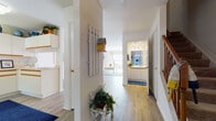 This two-story home features a welcoming entryway highlighted by light wood-look flooring that extends throughout the main level. The entry hall is accented with decorative wall hooks and charming coastal-inspired details, with a carpeted staircase fitted with a warm wood banister leading to the upper floor. The main level flows naturally from the entry into the living room, which is bathed in natural light from large windows and features a relaxed, coastal ambiance. Adjacent to the living area, a dining space benefits from a built-in pass-through window connecting directly to the kitchen, creating an open and functional layout. The kitchen is well-appointed with ample white cabinetry trimmed in warm wood tones, a skylight that floods the space with natural light, and sufficient counter space for meal preparation. A full bathroom on the main level features a single-sink vanity with white countertops and a shower-tub combination. Upstairs, the bedroom is spacious and serene, carpeted for comfort, with a large window providing ample natural light. Throughout the home, a consistent coastal color palette of white, navy, and warm wood tones creates a cohesive and inviting atmosphere.