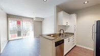 Located at 9710 Zelzah Ave in the Northridge neighborhood of Los Angeles, this single-floor residence offers a thoughtfully designed layout with a clean, modern aesthetic throughout. The open-concept kitchen is a standout feature, showcasing crisp white shaker cabinets with matte black hardware, granite countertops, a double-basin sink, and a full suite of stainless steel appliances including a gas range, microwave, dishwasher, and refrigerator. The kitchen flows seamlessly into the living area, which is finished with durable hardwood-style flooring that extends throughout the main living spaces, lending a warm and cohesive feel. Sliding glass doors in the living area open to a private balcony, allowing abundant natural light to fill the space. The bedrooms are carpeted for added comfort and are generously proportioned, with large windows dressed in vertical blinds that provide both light control and privacy. The primary bedroom connects to an en-suite bathroom featuring a wide vanity with a yellow stone countertop, white cabinetry, and a tiled walk-in shower with sliding glass doors. A second bathroom includes a shower-tub combination with a sleek sliding glass enclosure and hardwood-style flooring. The overall palette of neutral tones and clean finishes creates a bright, move-in ready environment.