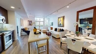 This thoughtfully designed single-floor residence offers a seamless blend of contemporary style and functional living. The open-concept main area unites the living, dining, and kitchen spaces, anchored by warm hardwood flooring that flows throughout. The living room is bright and inviting, featuring track lighting, large windows that welcome abundant natural light, and curated wall art that adds character to the crisp white walls. A generous sectional sofa arrangement centers the space, complemented by a round dining table nearby.

The kitchen is outfitted with rich dark cabinetry, light quartz countertops, a classic subway tile backsplash, and a full suite of stainless steel appliances including a gas range, microwave, dishwasher, and refrigerator. A sleek island with bar seating provides additional prep space and casual dining.

Both bedrooms are well-proportioned and bathed in natural light from large windows. Each offers a calm, restful atmosphere with clean white walls and hardwood floors. The primary bedroom features a warm wood headboard panel and bold artwork above.

Two bathrooms serve the residence — one a full bath finished in light tile with dark vanity cabinetry, and the other a compact half bath with a floating vanity and framed mirror. Curated artwork throughout gives the entire space a gallery-like quality.