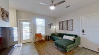 This single-floor residence showcases a clean, modern aesthetic defined by crisp white walls, light wood-style flooring, and an abundance of natural light throughout. The open-concept kitchen is a standout feature, equipped with stainless steel appliances including a refrigerator, range, and microwave, complemented by rich dark cabinetry, granite countertops, a subway tile backsplash, and a spacious island with seating. The kitchen flows directly into a dining area, creating a functional and social hub at the heart of the home. Two distinct living areas provide flexible use of space; one features a large flat-screen TV mounted on a white media console with natural wood accents and curated wall art, while the second offers a welcoming lounge with a ceiling fan, private outdoor access, and ample natural light from a large window. The primary bedroom is a serene retreat with soft carpet, warm lighting, and generous proportions. A second bedroom accommodates twin beds with ceiling fan and carpeted flooring, ideal for shared use. The bathroom is elegantly finished with a dual-sink granite vanity, dark wood cabinetry, and a soaking tub and shower adorned with large-format marble-look tile. Throughout, the design balances contemporary style with warm, livable comfort.