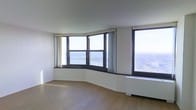 This single-floor high-rise space makes an immediate impression with its striking curved exterior window wall, which wraps gracefully around the main living area and frames breathtaking panoramic views of a wide river and the surrounding cityscape below. The abundance of floor-to-ceiling windows fitted with dark-framed casements and sheer curtains floods the interior with natural light throughout the day, creating a bright and airy atmosphere. The living area is finished with warm-toned hardwood flooring that complements the clean, white walls and adds a sense of understated elegance. A baseboard heating unit runs along the window wall, ensuring comfort in all seasons. The smooth, neutral-toned ceiling enhances the sense of openness, while the gently curving architectural lines of the window bay give the space a distinctive and sophisticated character that sets it apart from a conventional layout. The overall ambiance is calm and refined, with the ever-changing river and city views serving as a dynamic backdrop that brings life and energy to the interior. The space offers a seamless blend of architectural detail and natural scenery, making it a compelling and visually engaging environment.