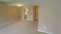This two-floor residential space offers a practical and comfortable layout designed for everyday living. The main living area is finished with wall-to-wall carpeting in a neutral beige tone, creating a warm and understated atmosphere throughout. Neutral-painted walls complement the soft flooring and contribute to the bright, airy feel of the space.

Adjacent to the living area is a compact kitchen, equipped with light wood cabinetry, a white electric range, and an over-the-range microwave. The kitchen features tile flooring, providing a clean contrast to the carpeted living space, and offers ample cabinet storage for functional use.

A hallway extends from the main living area, providing access to additional rooms beyond, including what appears to be a closet with bifold doors and further living spaces. The transition between the carpeted and tiled areas is seamless, reinforcing the cohesive flow of the floor plan.

The interior is illuminated by flush-mount ceiling lighting, ensuring consistent brightness throughout. Clean white baseboards and trim frame each room, adding a crisp, finished look to the overall design. The two-floor layout offers added versatility and separation of living spaces, making this a well-structured and adaptable home.