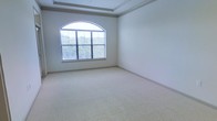 This single-story space presents a well-appointed room characterized by thoughtful architectural detailing and a bright, airy atmosphere. The room features a distinctive tray ceiling accented with crown molding, adding a sense of depth and elegance to the overall design. Soft, neutral-toned wall-to-wall carpeting covers the floor, contributing to a clean and comfortable environment.

A prominent arched window serves as the focal point of the room, fitted with horizontal blinds that allow for adjustable natural light control. The arch detail adds a classic architectural touch, while the window itself frames views of lush greenery outside, creating a pleasant connection to the outdoors. The generous influx of natural light enhances the room's open and welcoming feel.

The walls are finished in a crisp, neutral tone that complements the understated palette of the space. The room's clean lines, combined with the tray ceiling and arched window, suggest a refined yet versatile design suitable for a variety of uses. The overall ambiance is calm, bright, and polished, making it an adaptable and inviting space.