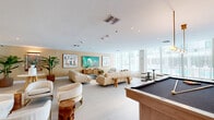 This elegantly designed amenity lounge offers a generous, open-plan layout spread across a single floor. The space is organized into several distinct functional zones, each thoughtfully appointed to encourage socializing and relaxation. Polished large-format terrazzo-style tile flooring flows seamlessly throughout, anchoring the bright, cohesive aesthetic.

A dedicated lounge area features plush, curved white bouclé armchairs with gold-accented bases arranged around organic wood side tables, complemented by a large sofa seating arrangement and a glass coffee table. Curated artwork and a large-format wall-mounted television add visual interest to the warm cream-toned walls.

A separate dining and booth section is defined by rich wood slat paneling and pendant globe lighting, with curved banquette seating in warm brown velvet surrounding round pedestal tables — evoking an upscale restaurant ambiance.

The space also includes a pool table, a bar-height kitchen island with warm wood cabinetry and botanical wallpaper accents, and high-top seating. Floor-to-ceiling windows along one wall flood the interior with natural light and provide direct views of the outdoor pool area, while sheer drapery softens the brightness. Lush tropical plants are placed throughout, enhancing the fresh, resort-inspired atmosphere.