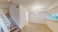 This two-story space is designed with a clean, neutral aesthetic throughout, featuring white walls, textured ceilings, and warm beige carpeting in the living and bedroom areas. The main level centers around a spacious living area anchored by a brick fireplace, lending a classic, cozy character to the room. A pendant light fixture overhead adds a decorative touch. Adjacent to the living area, an open pass-through connects to the kitchen, which is outfitted with white cabinetry, light countertops, tile flooring, and a full suite of white appliances including a refrigerator, electric range with overhead microwave, and dishwasher. A full bathroom on the main level features tile flooring, white cabinetry with ample under-sink storage, a built-in linen cabinet, a wide vanity mirror, and a toilet. A carpeted staircase with a white banister leads to the upper level, where carpeted rooms are served by ceiling fans with light fixtures and windows that allow natural light to filter in. The overall ambiance is bright, clean, and move-in ready, presenting a functional and well-organized layout across both floors.