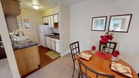This single-floor residence is designed with practicality and comfort in mind, offering a thoughtful layout that connects its key living spaces seamlessly. The kitchen is a galley-style space finished with white cabinetry accented by wood trim, dark granite-style countertops, a stainless steel double sink, and white appliances including a range, dishwasher, and refrigerator. Wood-look flooring runs through the kitchen, adding warmth and durability to the space.

Adjacent to the kitchen, the dining area accommodates a round table and chairs beneath a ceiling fan with lighting, creating an inviting spot for meals. This area flows naturally into the living room, which features plush carpeting, large windows with blinds that allow generous natural light, and a sliding glass door that adds additional brightness. The living room provides comfortable space for seating arrangements.

The primary bedroom is a well-proportioned carpeted retreat with windows on multiple walls, offering ample natural light and room for a full bedroom suite, with an attached bathroom nearby. A secondary bedroom features carpeting and a window, suitable for flexible use. The bathroom includes tile flooring, a vanity, toilet, and a tub-shower combination.