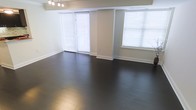 This single-floor space presents a clean, modern aesthetic defined by rich, dark hardwood flooring that spans the entire open area. The floors provide a striking contrast against the crisp white baseboards and light grey walls, creating a polished, contemporary look. Multiple large windows fitted with white horizontal blinds line the walls, allowing generous amounts of natural light to filter throughout the space while maintaining privacy. The windows also hint at outdoor views beyond. A structural column is integrated into the layout, subtly dividing the windowed wall without disrupting the overall openness of the room. The ceiling features a recessed beam detail and is equipped with a modern chandelier-style light fixture, adding a refined touch overhead. The neutral color palette of whites and greys throughout the walls and trim creates a versatile backdrop suitable for a variety of uses and furnishing styles. The overall ambiance is bright, airy, and well-maintained, with a minimalist character that emphasizes the quality of the finishes, particularly the deep-toned hardwood flooring that anchors the entire space.