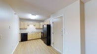 This single-floor space presents a straightforward, functional layout centered around a well-equipped kitchen. The kitchen features light wood cabinetry that lines the walls, providing ample storage space along both the upper and lower sections. The countertops are finished in a neutral tone, complementing the cabinetry and contributing to the room's clean, cohesive aesthetic. Black appliances, including a refrigerator, dishwasher, and range, offer a subtle contrast against the lighter cabinetry. A stainless steel sink is positioned along the counter, accompanied by under-cabinet lighting that enhances visibility during meal preparation. The flooring throughout is light wood-tone laminate, which adds warmth while remaining easy to maintain. Overhead, a flush-mounted light fixture provides consistent illumination across the space. The entry door, finished in a neutral tone with standard hardware, opens directly into this kitchen area, creating a practical and accessible entry point. The walls are painted in a crisp, neutral white, keeping the space feeling bright and open. Overall, the unit offers a clean, utilitarian design suited for everyday living.