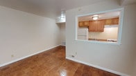 This single-floor space offers a straightforward and functional layout designed for comfortable everyday living. The main living area is anchored by classic parquet wood flooring in a warm honey tone, adding character and a timeless quality to the space. Crisp white walls and flush-mount ceiling lighting keep the area feeling bright and well-lit throughout.

A defining architectural feature is the pass-through window that connects the living area to the kitchen, creating an open sightline and an informal sense of connectivity between the two spaces. The kitchen is equipped with warm-toned oak cabinetry providing ample storage, a white refrigerator, and a sink with a granite-style countertop. The kitchen is finished with matching flush-mount lighting and light-colored walls that complement the cabinetry.

The living area includes a baseboard heating unit along the wall, ensuring year-round comfort. Subtle architectural details such as clean white baseboards and a recessed soffit area add dimension to the otherwise streamlined interior. Floor vents are thoughtfully integrated into the flooring. The overall ambiance of the space is clean, neutral, and move-in ready, offering a solid foundation for personalization.