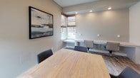This business center is designed to provide a functional and comfortable workspace within a single, efficiently organized room. At the heart of the space sits a solid wood conference table surrounded by upholstered dark chairs, making it well-suited for meetings or collaborative work sessions. Natural light filters in through multiple windows dressed with woven wood Roman shades, offering a warm, refined aesthetic while maintaining privacy. A standout feature is the built-in granite countertop workstation that wraps along one wall, accompanied by ergonomic rolling chairs — ideal for individual focused work or computer tasks. Recessed ceiling lighting supplements the natural light, ensuring the space remains bright and well-illuminated throughout the day. The neutral warm-gray wall tones are complemented by a large framed abstract artwork, lending a touch of sophistication to the professional setting. Durable wood-look flooring transitions seamlessly throughout, contributing to the clean, contemporary design. A glass-paneled door provides easy access while maintaining an open, connected feel to the surrounding area. The overall ambiance strikes a balance between polished professionalism and approachable comfort, making this business center a versatile resource for productivity.