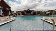 Located at 4627–4633 Monroe Way in Fredericksburg, Virginia, this single-story community clubhouse serves as a vibrant social hub designed with both comfort and recreation in mind. The building's classic brick exterior with white trim columns provides an inviting first impression, complemented by a sprawling resort-style swimming pool surrounded by a spacious patio deck outfitted with lounge chairs and shade umbrellas.

Inside, the clubhouse immediately impresses with dramatically soaring two-story ceilings that flood the interior with natural light through tall clerestory windows. The main lounge area features rich hardwood-style flooring, a sleek wall-mounted fireplace, a flat-screen TV, and boldly styled furniture in vibrant teal and orange tones that give the space a modern, energetic character.

The gathering and game area offers high-top seating around bistro-style tables alongside a full-size shuffleboard table, making it ideal for socializing and recreation. Artwork accents the walls, adding color and visual interest throughout. Large windows dressed with floor-to-ceiling curtains draw in ample sunlight while offering views of the surrounding community. A dedicated exit door provides direct access to the pool deck, seamlessly connecting the indoor and outdoor amenity spaces. Overall, the clubhouse delivers a polished and welcoming environment for residents to gather, relax, and enjoy.
