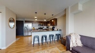 This single-floor residence is thoughtfully designed with an open-concept layout that seamlessly connects the kitchen, dining, and living areas. The kitchen is a standout feature, showcasing rich dark-brown cabinetry with ample upper and lower storage, granite countertops, and a full suite of stainless steel appliances, including a side-by-side refrigerator with water dispenser, electric range, microwave, and dishwasher. A peninsula with a granite overhang accommodates four metal bar stools, providing a casual dining and gathering space. Three pendant lights hang above, adding a modern touch to the kitchen's warm aesthetic.

The living room offers a comfortable, relaxed atmosphere with light hardwood-style flooring carried throughout the main areas, a large sectional sofa, a wooden coffee table, and a window that brings in natural light. The bedroom features soft carpeting, a window with outdoor views, and a clean, serene design. The bathroom presents wood-toned cabinetry, a white vanity, and hardwood-style flooring. Neutral wall tones and a textured ceiling are consistent throughout, creating a cohesive and welcoming environment across all rooms.