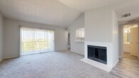 This single-floor residence offers a thoughtfully laid-out interior with a blend of practical features and comfortable living spaces. The living room serves as the centerpiece of the home, distinguished by soaring vaulted ceilings that lend an open, airy feel to the space. A classic fireplace with glass doors anchors one wall, while a sliding glass door fitted with vertical blinds opens to an outdoor area, allowing natural light to flood the room. Soft, neutral carpet extends throughout the living areas and bedrooms, contributing to a warm and cohesive aesthetic. A pass-through opening connects the living room to the kitchen, which is equipped with white shaker-style cabinetry, light stone-look countertops, and a stainless steel dishwasher. The bedrooms are generously proportioned, each featuring plush carpet, natural light from windows with blinds, and closet storage. The bathroom includes a tiled shower-tub combination, wood-look tile flooring, and a white vanity. A dedicated laundry area accommodates a full-size side-by-side washer and dryer, with overhead shelving for additional storage. The overall color palette of soft whites and grays creates a clean, neutral canvas throughout the home.