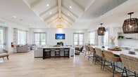 Located in Surf City, NC, this single-floor coastal clubhouse at 1040 Vinson Way is a beautifully designed communal space that blends coastal charm with modern sophistication. The open-concept layout is anchored by a dramatic vaulted ceiling with exposed white-painted beams and shiplap detailing, creating an airy, expansive atmosphere from the moment you enter.

Light-toned hardwood flooring runs throughout, complementing the crisp white walls and abundant natural light that pours in through multiple sets of French doors and large windows. The main gathering area features a generous lounge space centered around a tiled fireplace with a wall-mounted television above, creating a welcoming focal point flanked by comfortable seating.

Adjacent to the lounge, a large kitchen island with a white quartz countertop is surrounded by woven rattan barstools, while a well-appointed cabinetry run in sage green offers open wood shelving and a beverage cooler beneath. Pendant lights in woven and rattan finishes add to the coastal aesthetic throughout. A secondary seating nook provides additional intimate lounge space with casual furniture and coastal-themed artwork. Recessed lighting and carefully chosen pendant fixtures ensure the space is warmly lit at all times, making this clubhouse an inviting retreat for gatherings and everyday use alike.