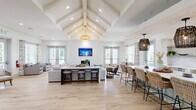 Located in Surf City, NC, this single-floor coastal clubhouse at 1040 Vinson Way is a beautifully designed communal space that blends coastal charm with modern sophistication. The open-concept layout is anchored by a dramatic vaulted ceiling with exposed white-painted beams and shiplap detailing, creating an airy, expansive atmosphere from the moment you enter.

Light-toned hardwood flooring runs throughout, complementing the crisp white walls and abundant natural light that pours in through multiple sets of French doors and large windows. The main gathering area features a generous lounge space centered around a tiled fireplace with a wall-mounted television above, creating a welcoming focal point flanked by comfortable seating.

Adjacent to the lounge, a large kitchen island with a white quartz countertop is surrounded by woven rattan barstools, while a well-appointed cabinetry run in sage green offers open wood shelving and a beverage cooler beneath. Pendant lights in woven and rattan finishes add to the coastal aesthetic throughout. A secondary seating nook provides additional intimate lounge space with casual furniture and coastal-themed artwork. Recessed lighting and carefully chosen pendant fixtures ensure the space is warmly lit at all times, making this clubhouse an inviting retreat for gatherings and everyday use alike.