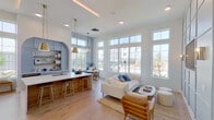 This thoughtfully designed single-floor space showcases a bright, open-concept layout that seamlessly blends social, recreational, and lounge areas. Throughout the interior, light-toned hardwood flooring, crisp white walls, and soft blue-grey accents create a cohesive, contemporary aesthetic. Soaring ceilings with recessed lighting and large floor-to-ceiling windows flood every corner with natural light and offer views of the surrounding community.

The main lounge area features a spacious seating arrangement with a white sectional sofa, anchored by a geometric-patterned rug and wall-mounted displays on a paneled accent wall. Warm wood slatted dividers elegantly separate functional zones without sacrificing openness.

A fully appointed kitchen and bar area includes a substantial island with a white quartz countertop, wood-paneled base, and bar seating. Gold pendant lighting and a charming built-in arched shelving nook add distinctive character. Adjacent to the kitchen, a cozy dining nook with a built-in cushioned bench, round table, and statement pendant light provides an intimate gathering spot framed by tall windows.

The expansive recreation zone accommodates a pool table alongside additional casual seating, while a separate area features arcade-style entertainment and lounge chairs. The transition zones include hexagonal tile flooring details and natural wood beam accents, adding warmth and textural variety throughout this well-appointed community space.