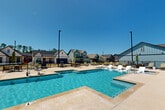 Located at 180 Hughes Rd in Madison, Alabama, this outdoor community amenity area centers around a generously sized resort-style swimming pool designed for both relaxation and recreation. The pool features a multi-section layout, including a main swimming area and a shallow wading shelf with in-water lounge chairs, ideal for unwinding in the Alabama sun. The surrounding pool deck is finished with a smooth, light-toned concrete surface that stays comfortable underfoot and provides ample space for lounging. Numerous chaise lounge chairs are arranged throughout the deck, offering plenty of seating options for residents. Outdoor dining tables with umbrellas are also situated around the perimeter, creating casual gathering spots. The area is enclosed by a black iron fence, ensuring safety and privacy while maintaining open sightlines to the surrounding residential neighborhood. Tall light poles are positioned throughout the space, extending the usability of the pool area into the evening hours. A community building with a covered porch is visible along one edge of the pool deck, adding a social hub to the amenity space. The backdrop of craftsman and farmhouse-style homes contributes to the charming, neighborhood-oriented atmosphere of this well-appointed outdoor retreat.