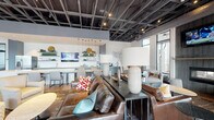 Located at 7740 Midtown Market Drive in Raleigh, NC, this single-floor community clubhouse combines modern industrial aesthetics with warm, inviting interiors. The open-plan layout is anchored by an exposed structural ceiling with dark-painted ductwork and grid framework, lending a loft-like character to the space. Rich hardwood flooring flows throughout, complementing the varied seating zones designed for both relaxed socializing and focused gathering.

The lounge area features plush leather sofas accented with colorful throw pillows, arranged around a tiled fireplace flanked by a wall-mounted TV — an ideal spot for casual conversation. Adjacent seating vignettes offer additional comfort with upholstered armchairs and side tables. A bold built-in banquette with magenta upholstery and curated wall art adds a vibrant, gallery-like focal point to one section of the room.

The full kitchen is well-appointed with stainless steel appliances, dark cabinetry, a central island with a light wood countertop, open display shelving, a decorative tile backsplash, and pendant lighting featuring ring-and-Edison-bulb fixtures. A dedicated dining table provides a communal workspace or gathering area. Floor-to-ceiling windows along the perimeter flood the space with natural light and offer views of the surrounding Midtown neighborhood, creating a bright and welcoming atmosphere throughout.