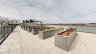 Located at 2812 Rue Ontario E in Montréal's vibrant Hochelaga-Maisonneuve neighbourhood, this shared rooftop terrace is a thoughtfully designed communal outdoor space offering a wide range of amenities for residents. The terrace is finished with large-format light grey porcelain tiles throughout, lending a clean, contemporary aesthetic to the entire level.

At the heart of the space is a series of numbered raised garden planters constructed in sleek grey metal, arranged in organized rows across the terrace. These individual plots provide residents with dedicated space for urban gardening and greenery. Ornamental grasses and seasonal plantings add texture and life to the landscape.

A standout feature is the covered pergola structure, fitted with string lighting and furnished with warm-toned wooden dining tables and benches, creating an inviting outdoor gathering area. Adjacent to this is a fully equipped outdoor kitchen and BBQ station with dark cabinetry, ideal for communal cooking and entertaining.

The terrace offers unobstructed panoramic views of the Montréal skyline, including a distant sightline toward Mont-Royal, providing a compelling urban backdrop. The surrounding building façade in light brick with grey balconies frames the interior courtyard portions of the rooftop, reinforcing the modern architectural character of the development.